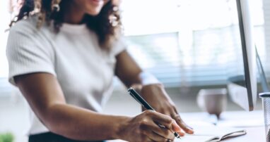 A person writes in a notebook at a computer desk