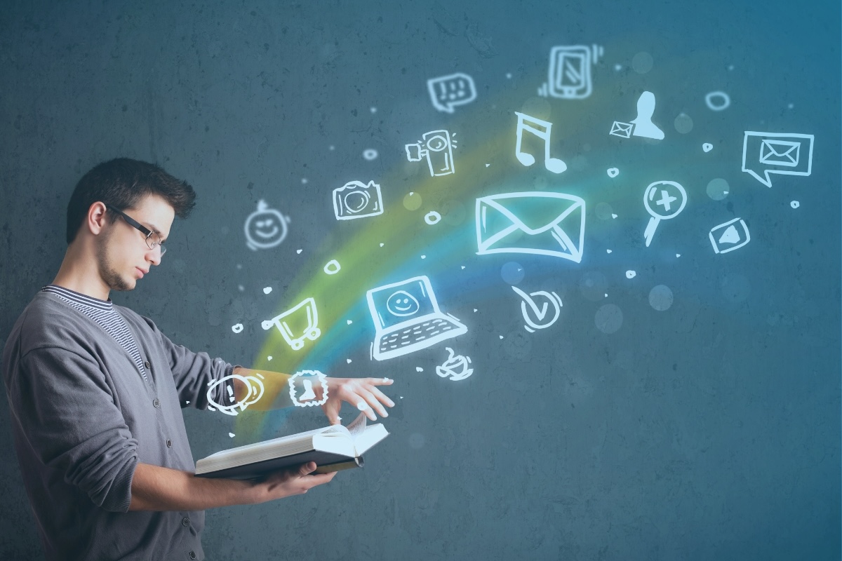 A person turning a page in a book that is spilling out images of laptops, email icons, and emojis.