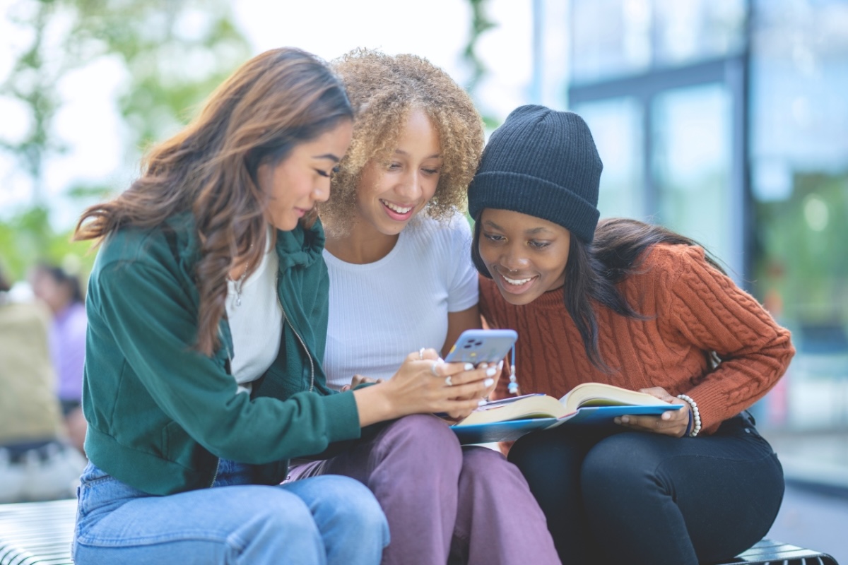 Three smiling people look at a smartphone together outdoors.
