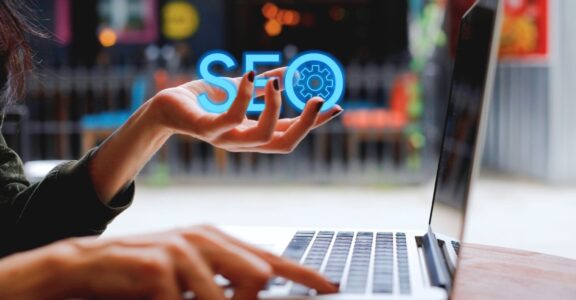 A person presses a key on a laptop with one hand and holds the word "SEO" with the other hand