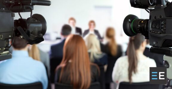 A large group of people, some with cameras, facing two people seated at a table